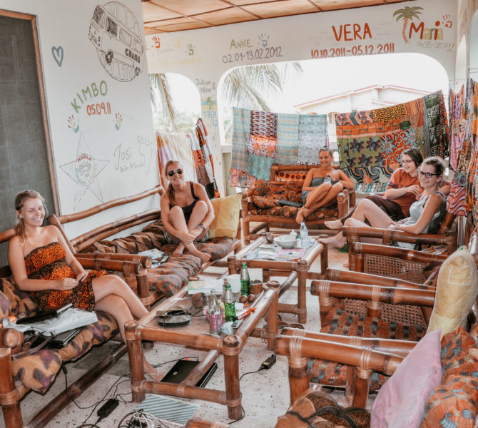 Gemütliche Lounge mit Menschen in Sommerkleidung im wayers-home, umgeben von bunten Stoffen und Wandmalereien in entspannter, tropischer Atmosphäre.