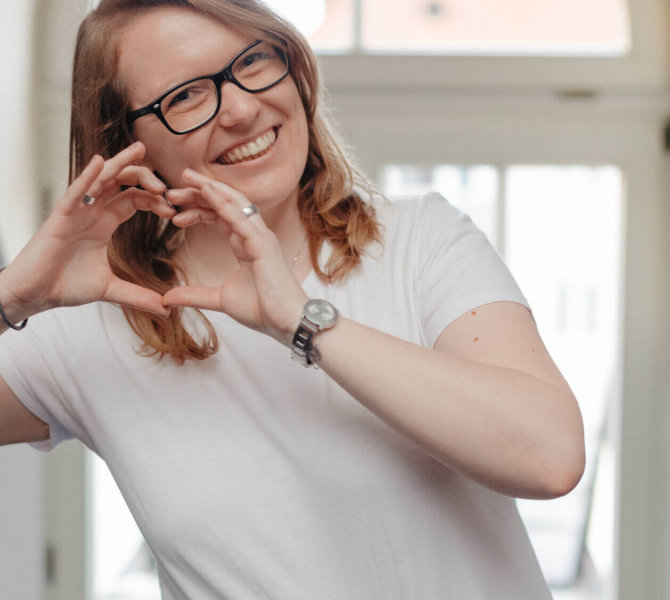 Eine lächelnde Frau formt mit den Händen ein Herz, steht vor einem hellen Fenster und trägt Brille sowie ein weißes T-Shirt.