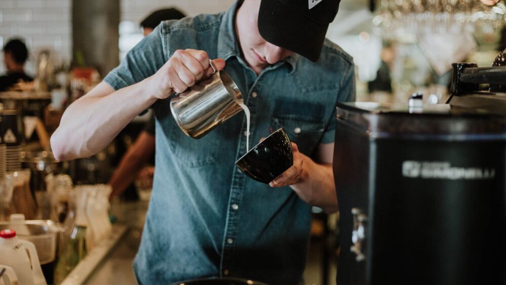 Ein Barista gießt konzentriert Milchschaum in eine Tasse, um Latte Art zu kreieren, in einer geschäftigen Café-Atmosphäre.