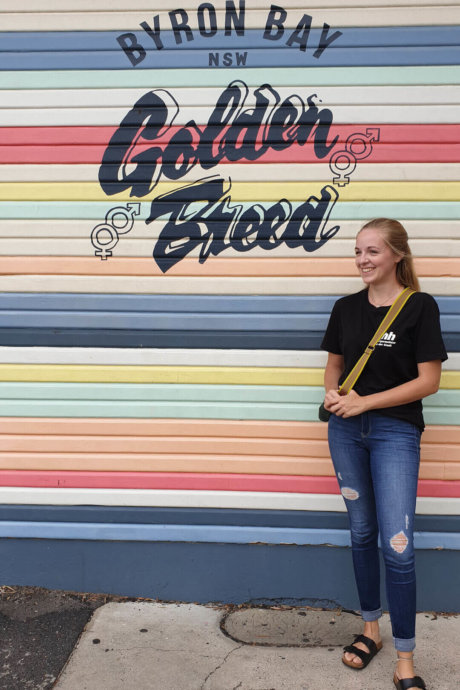 Eine junge Frau steht lächelnd vor einer bunten Wand mit der Aufschrift „Byron Bay NSW Golden Breed“.