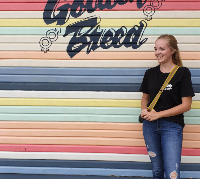 Eine junge Frau steht lächelnd vor einer bunten Wand mit der Aufschrift „Byron Bay NSW Golden Breed“.