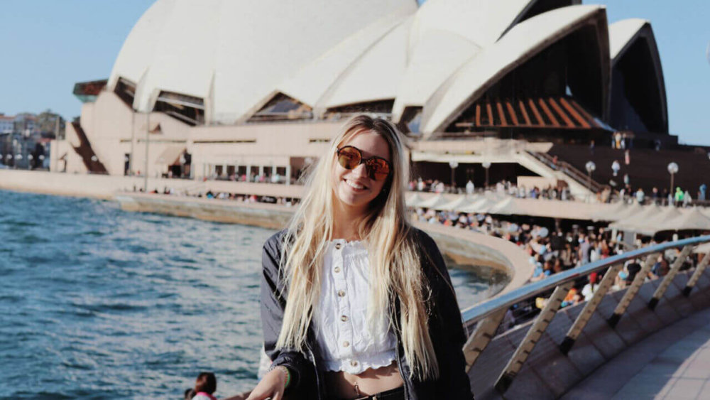 Eine Frau posiert lächelnd vor dem Sydney Opera House an einem sonnigen Tag mit Blick auf das Wasser und die Promenade.