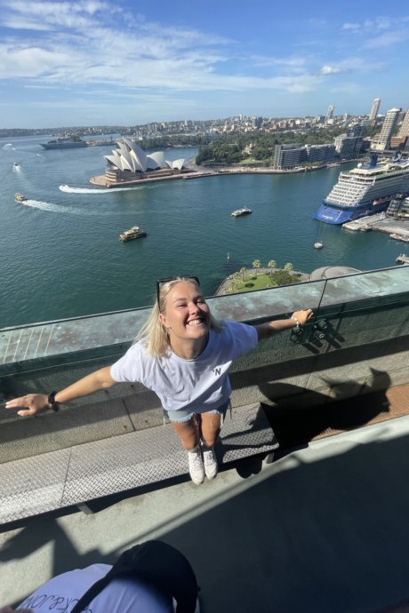 Eine Frau steht fröhlich auf der Sydney Harbour Bridge mit Blick auf das Opernhaus und den Hafen bei sonnigem Wetter.
