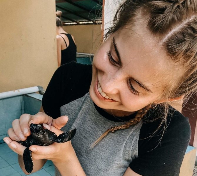 Junge Frau hält lächelnd eine kleine Meeresschildkröte in der Hand bei einem Schutzprojekt für Meeresschildkröten auf Bali