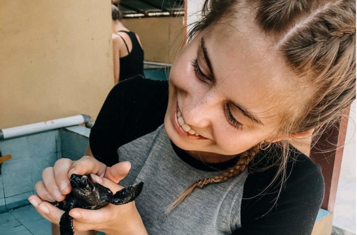 Junge Frau hält lächelnd eine kleine Meeresschildkröte in der Hand bei einem Schutzprojekt für Meeresschildkröten auf Bali