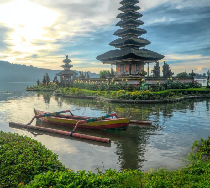 Der Ulun Danu Tempel auf Bali spiegelt sich im Wasser des Bratan Sees mit traditionellem Boot im Vordergrund und dramatischem Himmel bei Sonnenuntergang.