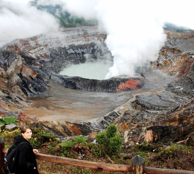Frau blickt auf dampfenden Krater eines aktiven Vulkans mit Schwefelsee umgeben von rauer Lavalandschaft und dichter Vegetation.