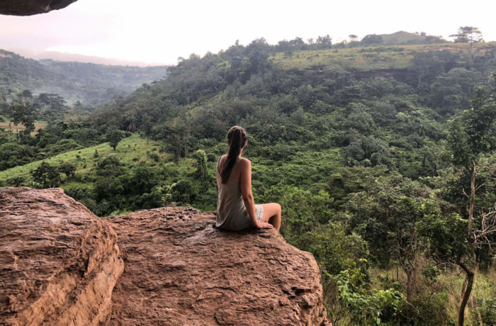 Frau sitzt auf dem Umbrella Rock in Ghana und blickt über eine grüne hügelige Landschaft mit dichter Vegetation bei weichem Abendlicht