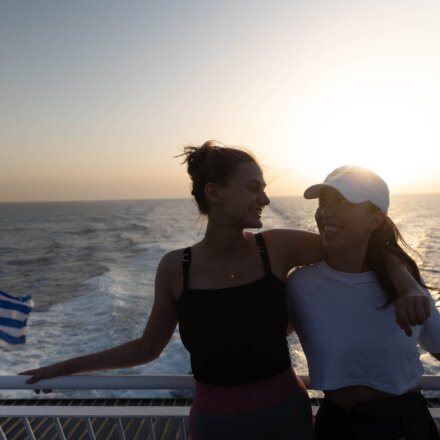 Zwei junge Frauen lachen Arm in Arm auf einer Fähre bei Sonnenuntergang mit Blick auf das Meer und griechische Flagge.