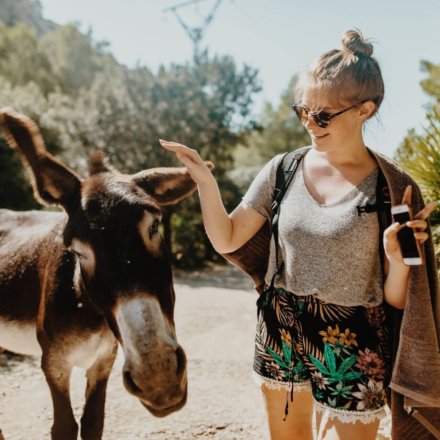 Junge Frau trifft auf einem sonnigen Wanderweg auf einen Esel und streckt ihm freundlich die Hand entgegen in Griechenland