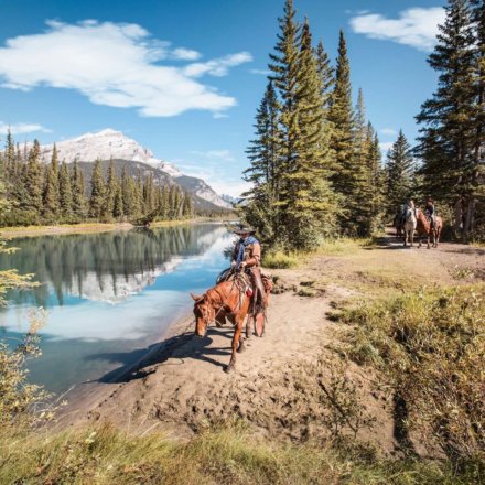 Reiter auf Pferden am Flussufer umgeben von Nadelbäumen und Bergen bei klarem Himmel während eines Farmstays in Kanada.