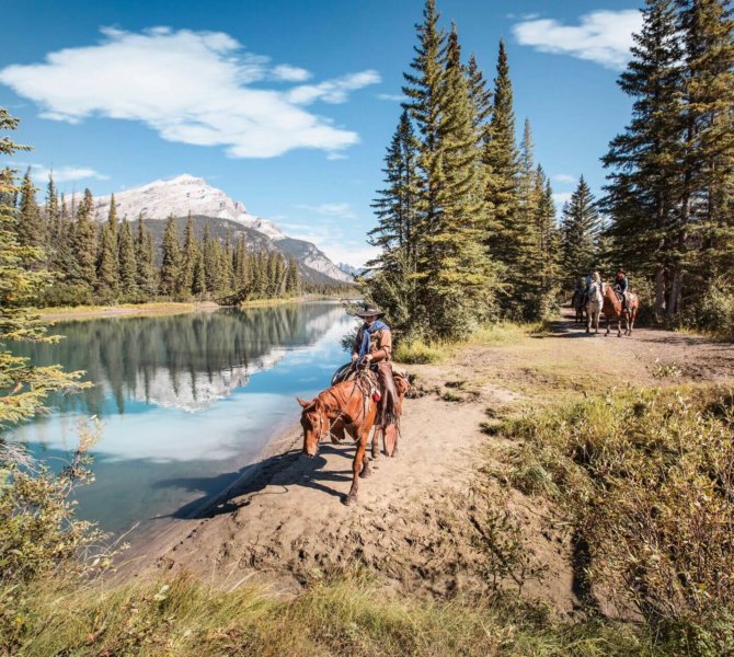 Reiter auf Pferden am Flussufer umgeben von Nadelbäumen und Bergen bei klarem Himmel während eines Farmstays in Kanada.