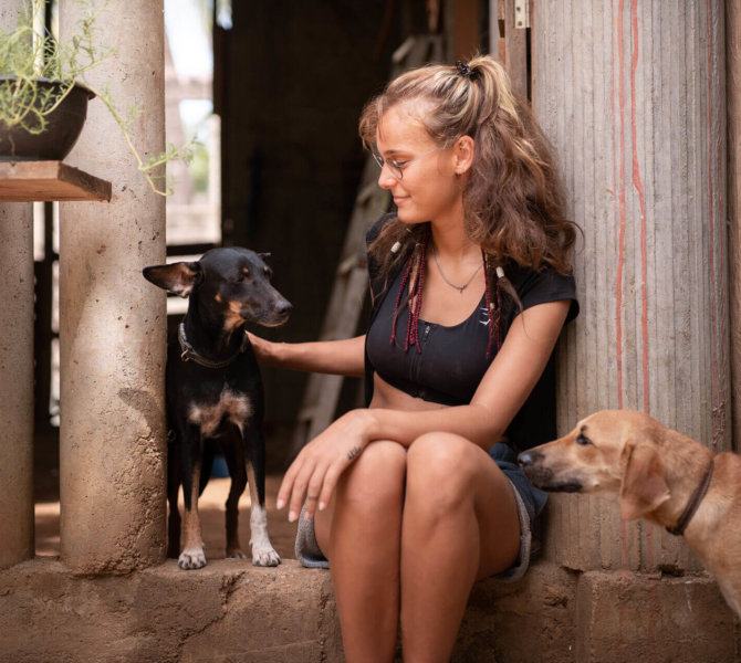 Junge Frau sitzt in einem offenen Gebäude und streichelt einen Hund während ein zweiter Hund neugierig danebensteht.