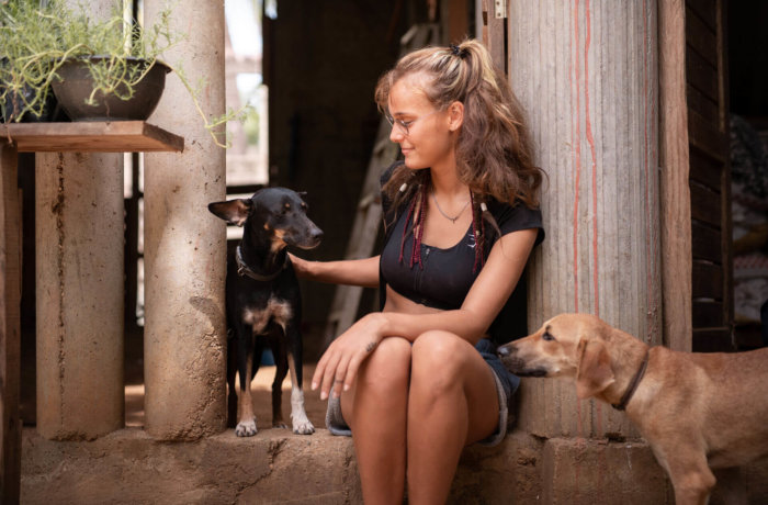 Junge Frau sitzt in einem offenen Gebäude und streichelt einen Hund während ein zweiter Hund neugierig danebensteht.