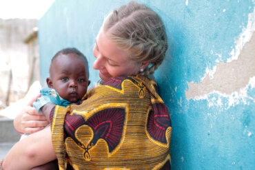 Freiwillige in bunter Kleidung in Sansibar hält ein Baby liebevoll im Arm vor blauer, verwitterter Wand und lächelt es an