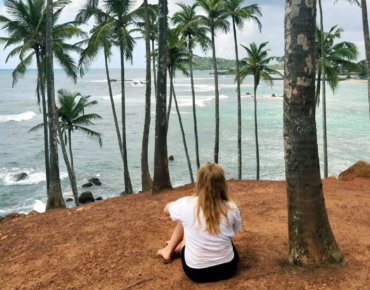 Frau sitzt auf Klippe unter Palmen und blickt auf das Meer während einer Reise auf Sri Lanke