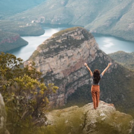 Frau steht mit erhobenen Armen auf Felsvorsprung mit Blick auf beeindruckende Berglandschaft und Fluss in weiter Ferne in Südafrika