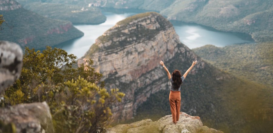 Frau steht mit erhobenen Armen auf Felsvorsprung mit Blick auf beeindruckende Berglandschaft und Fluss in weiter Ferne in Südafrika
