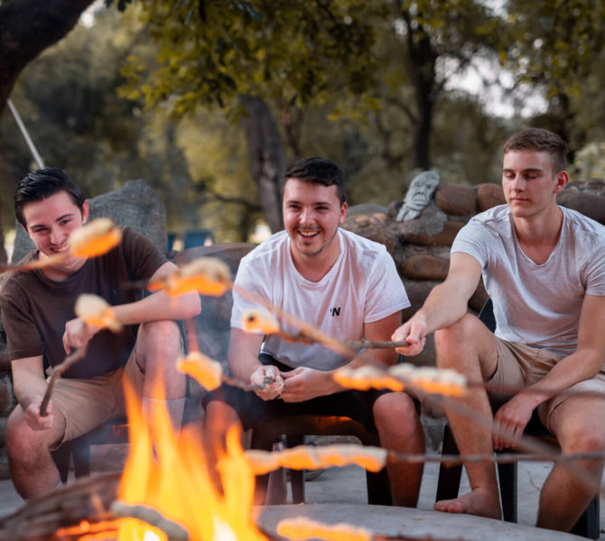 Gruppe junger Männer sitzt entspannt am Lagerfeuer und grillt Brot inmitten der Natur beim Wildlifeprojekt im Krüger Nationalpark in Südafrika