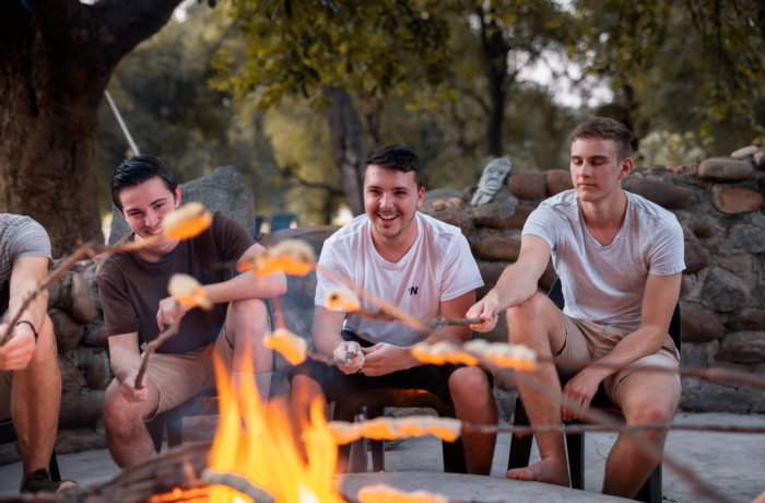 Gruppe junger Männer sitzt entspannt am Lagerfeuer und grillt Brot inmitten der Natur beim Wildlifeprojekt im Krüger Nationalpark in Südafrika