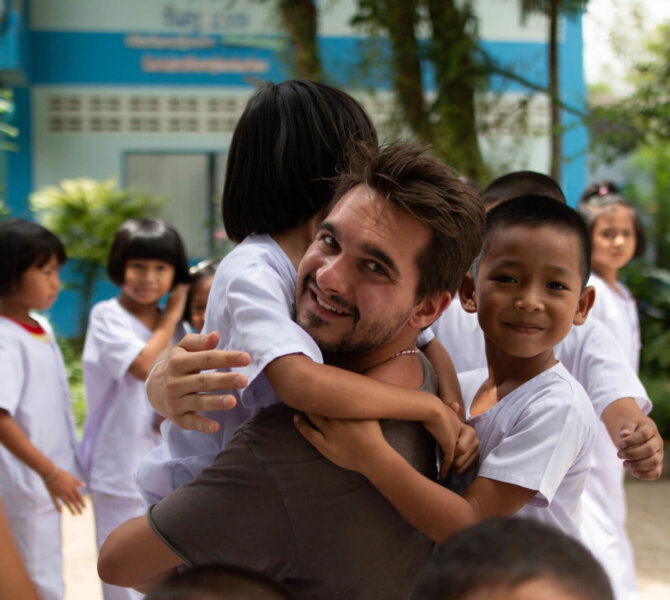 Mann wird lachend von Kindern in Schuluniform umarmt auf einem Schulhof in Thailand bei der Freiwilligenarbeit