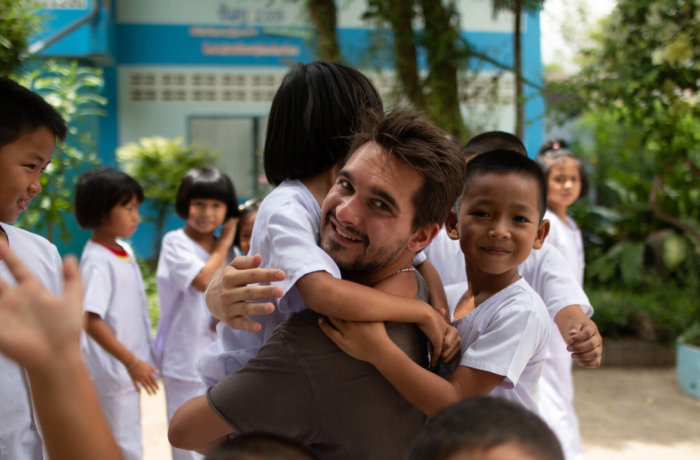 Mann wird lachend von Kindern in Schuluniform umarmt auf einem Schulhof in Thailand bei der Freiwilligenarbeit