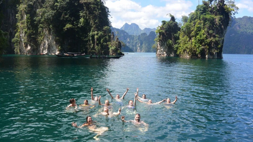 Gruppe junger Menschen schwimmt lachend im türkisfarbenen See, umgeben von steilen Felsen und tropischer Naturkulisse in Thailand.