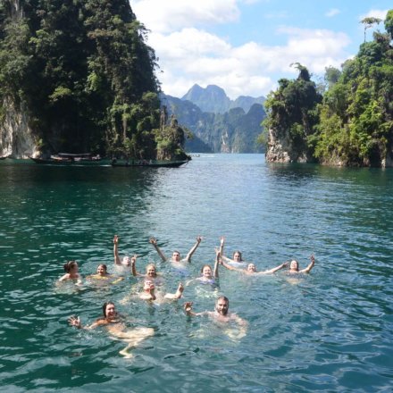 Gruppe junger Menschen schwimmt lachend im türkisfarbenen See, umgeben von steilen Felsen und tropischer Naturkulisse in Thailand.